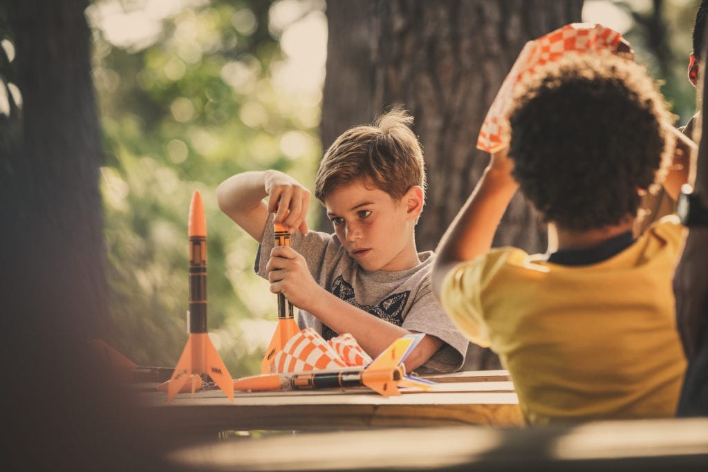 Two boys build rockets outside at a wooden picnic table, learning STEM skills.